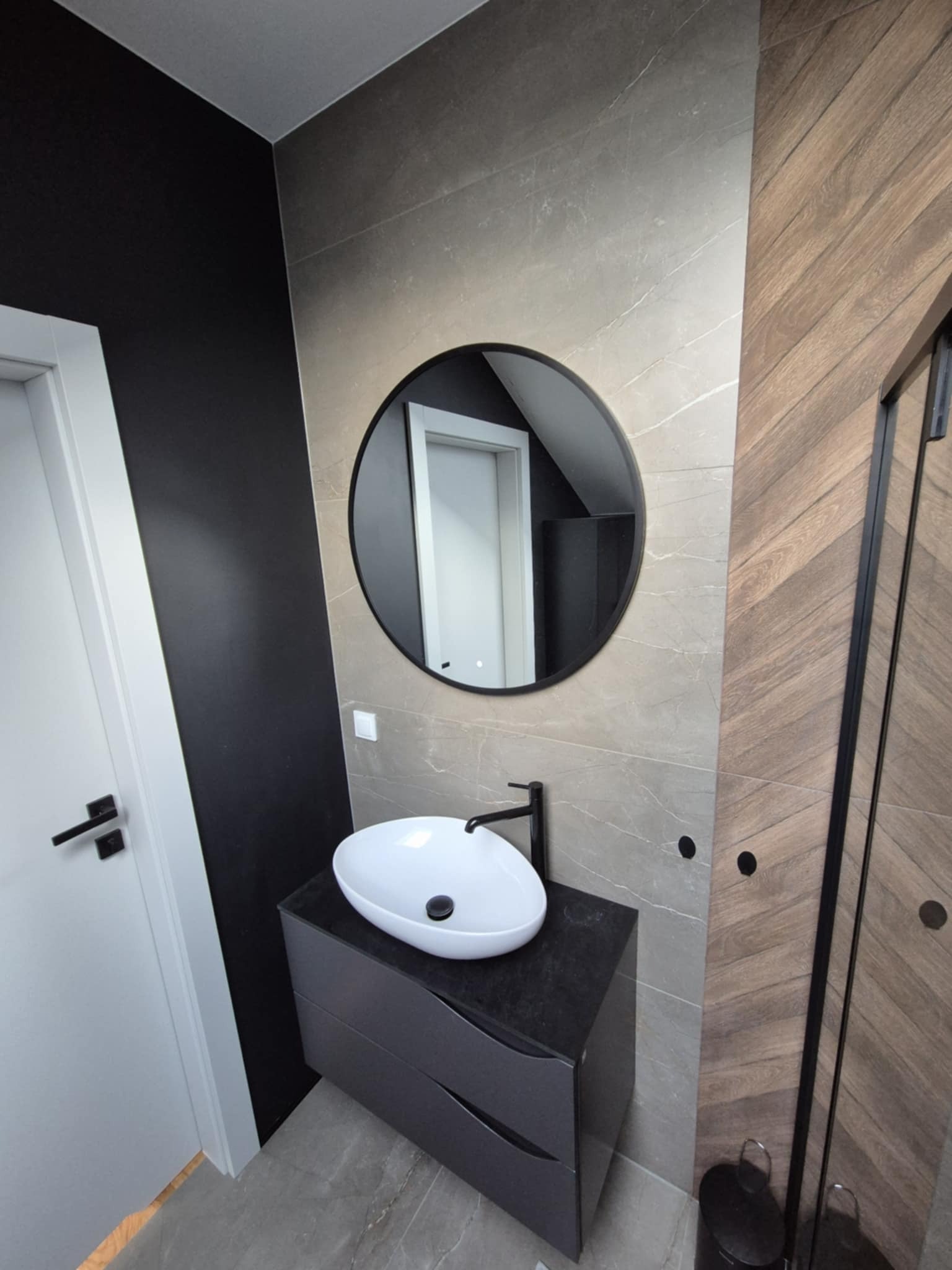 Nowoczesna łazienka z owalną umywalką nablatową, okrągłym lustrem i kabiną prysznicową z czarnymi profilami. Ściany wykończone płytkami imitującymi marmur i drewno.