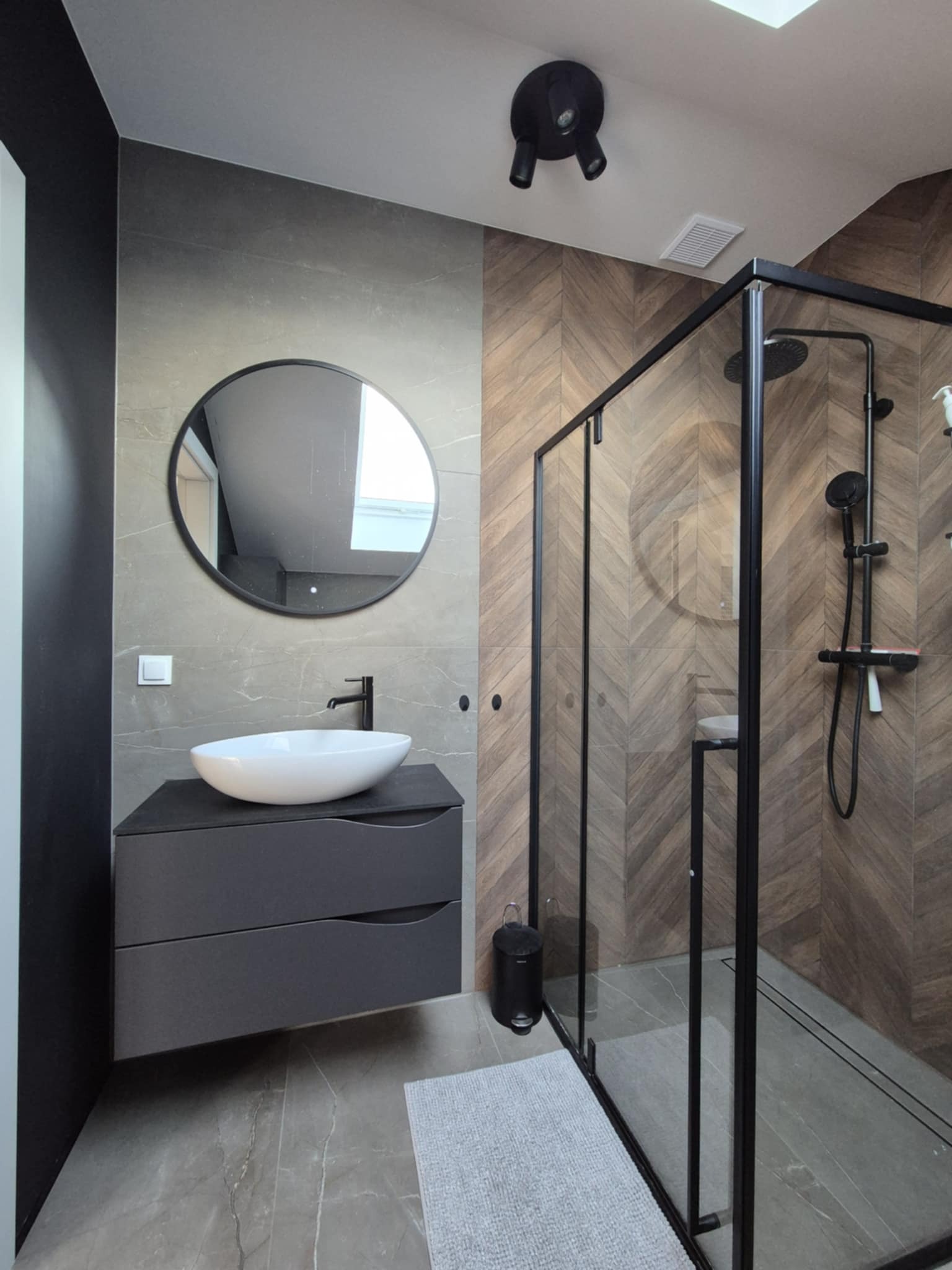 Nowoczesna łazienka z kabiną prysznicową typu walk-in, umywalką nablatową na szarej szafce i okrągłym lustrem. Ściany wyłożone płytkami w odcieniach szarości i brązu.