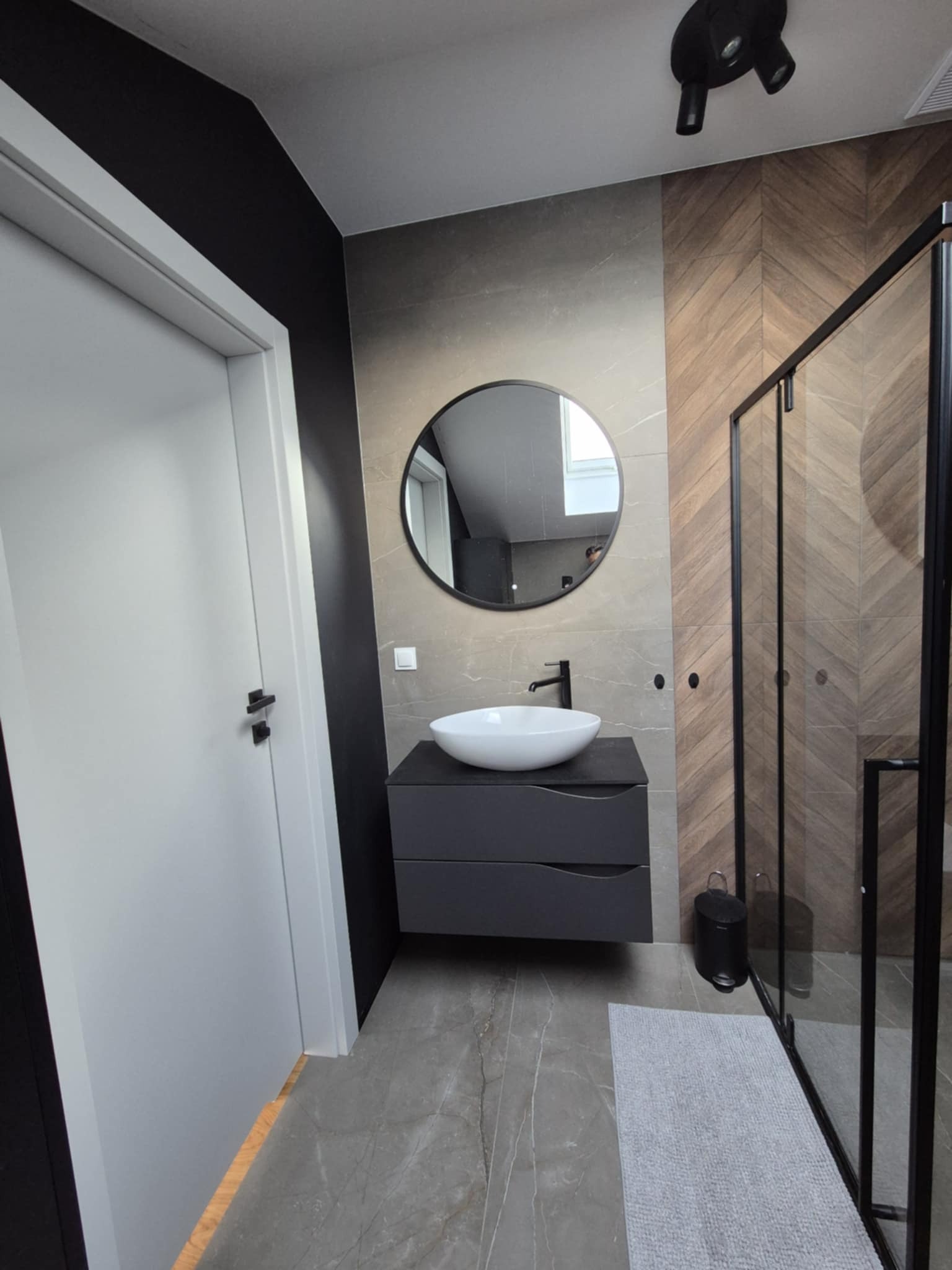 Nowoczesna łazienka z okrągłym lustrem, umywalką nablatową i kabiną prysznicową z czarnymi profilami. Na ścianie płytki imitujące drewno ułożone w jodełkę.