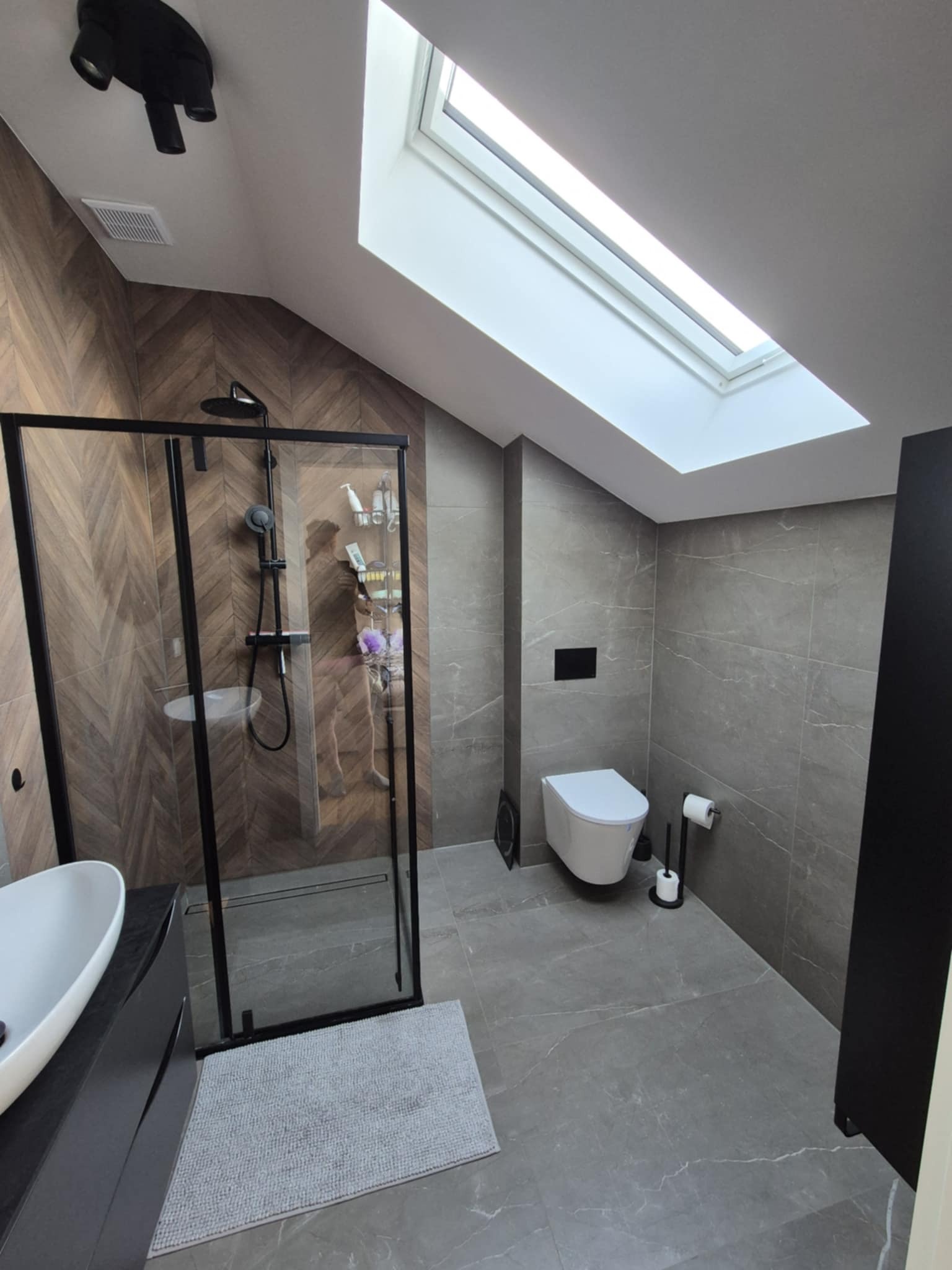 Nowoczesna łazienka na poddaszu z kabiną prysznicową typu walk-in, umywalką nablatową i toaletą. Uwagę zwracają płytki imitujące drewno w strefie prysznica i okno dachowe.