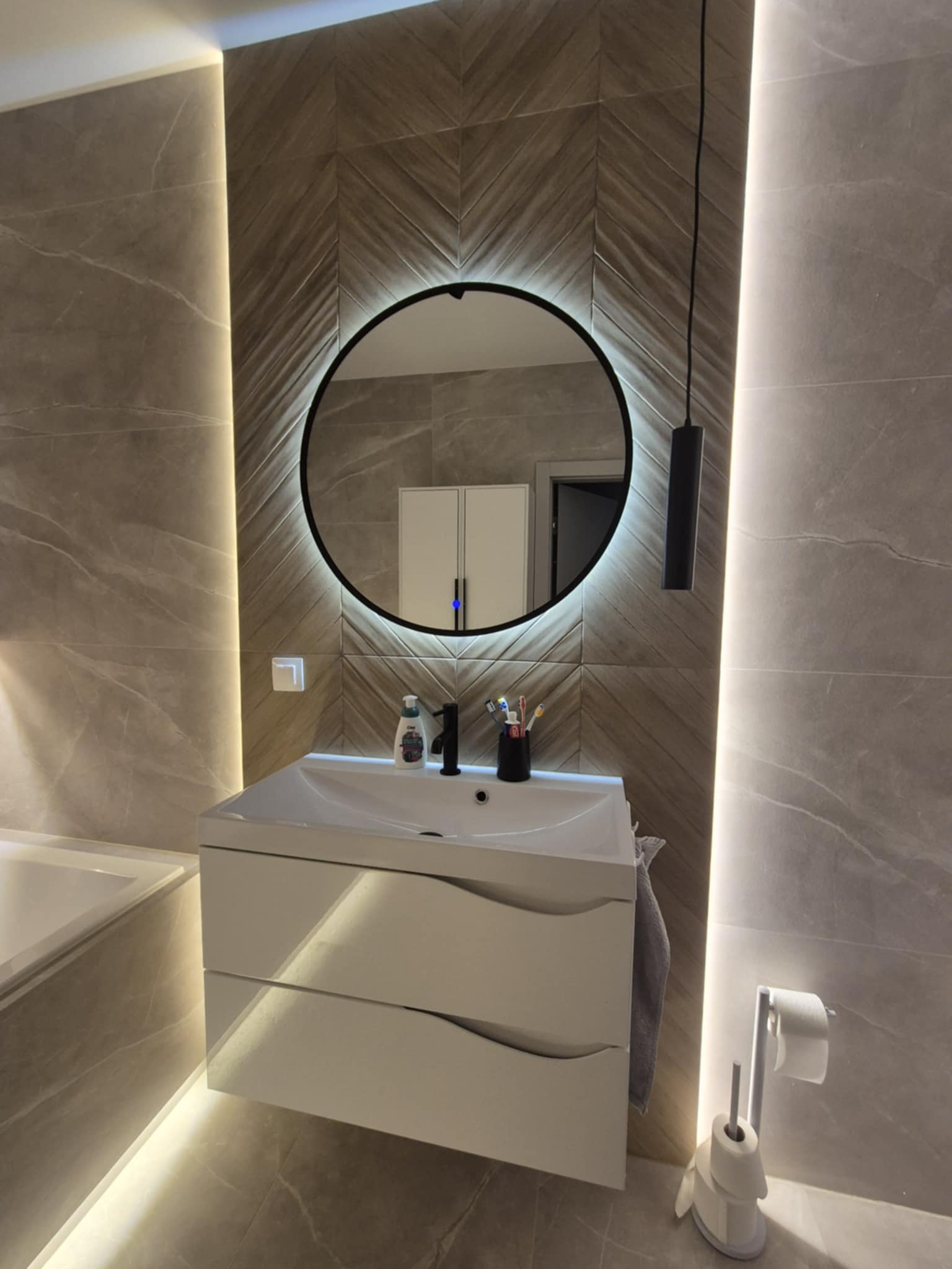 Nowoczesna łazienka z umywalką na szafce, okrągłym lustrem z podświetleniem LED i płytkami w odcieniach szarości ułożonymi w jodełkę na ścianie.