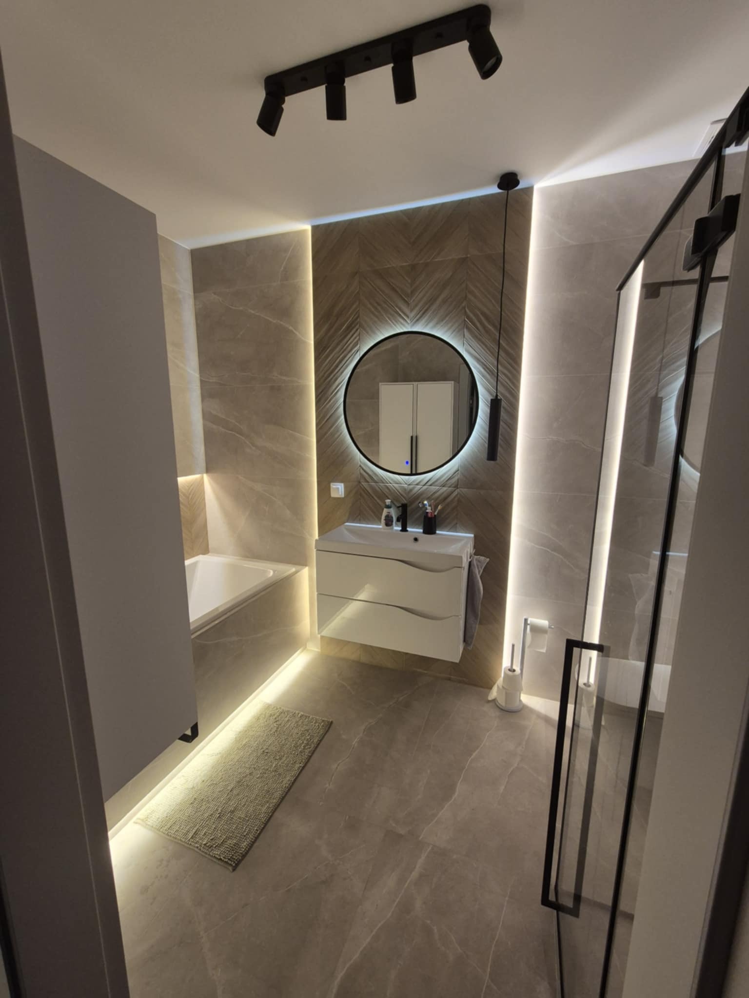 Nowoczesna łazienka z okrągłym lustrem podświetlanym LED, beżowe płytki imitujące marmur, kabina prysznicowa z czarnymi ramami i punktowe oświetlenie sufitowe.