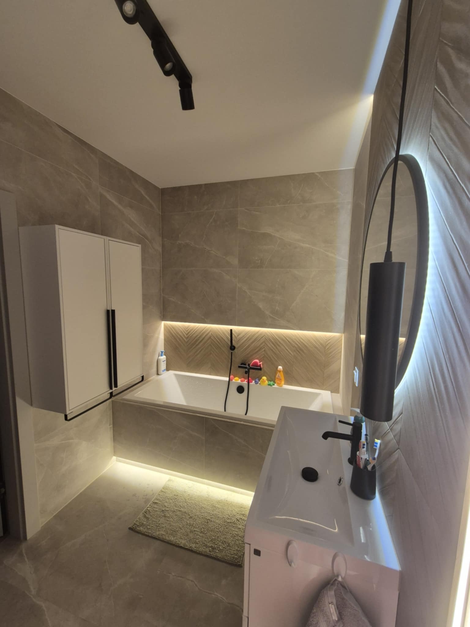 Nowoczesna łazienka z podświetleniem LED, marmurowymi płytkami i owalnym lustrem. Umywalka z szafką, wanna z baterią i wisząca szafka tworzą spójną całość.