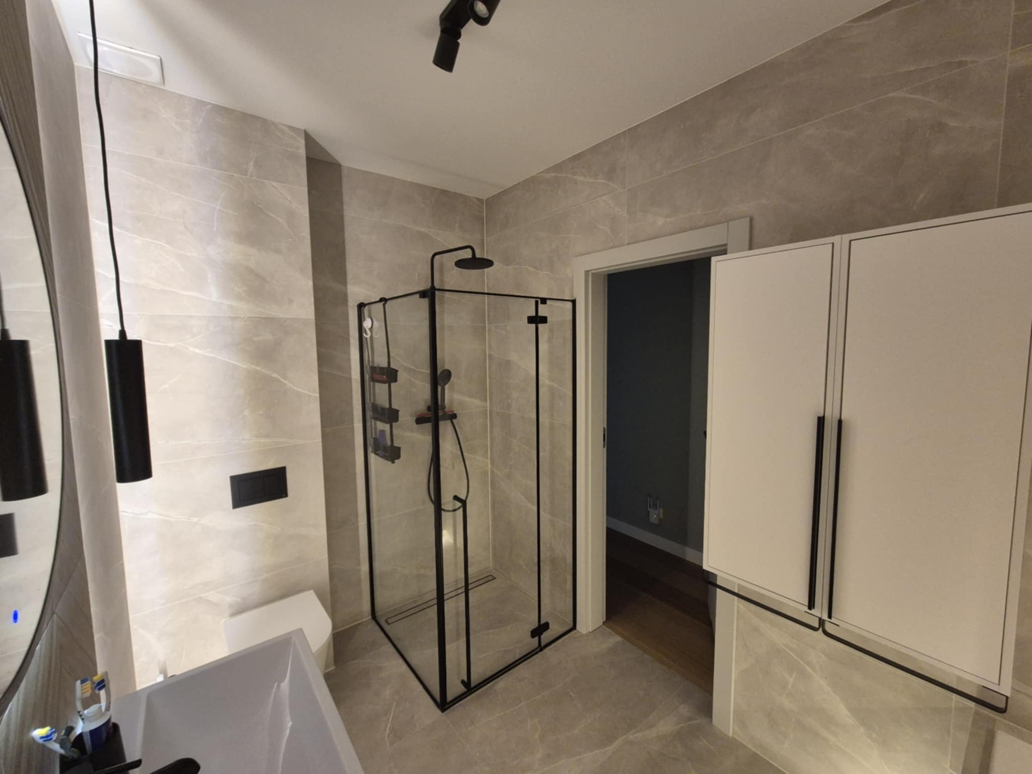 Nowoczesna łazienka w beżowych płytkach z kabiną prysznicową typu walk-in z czarnymi profilami, białą szafką i elementami armatury w kolorze czarnym.