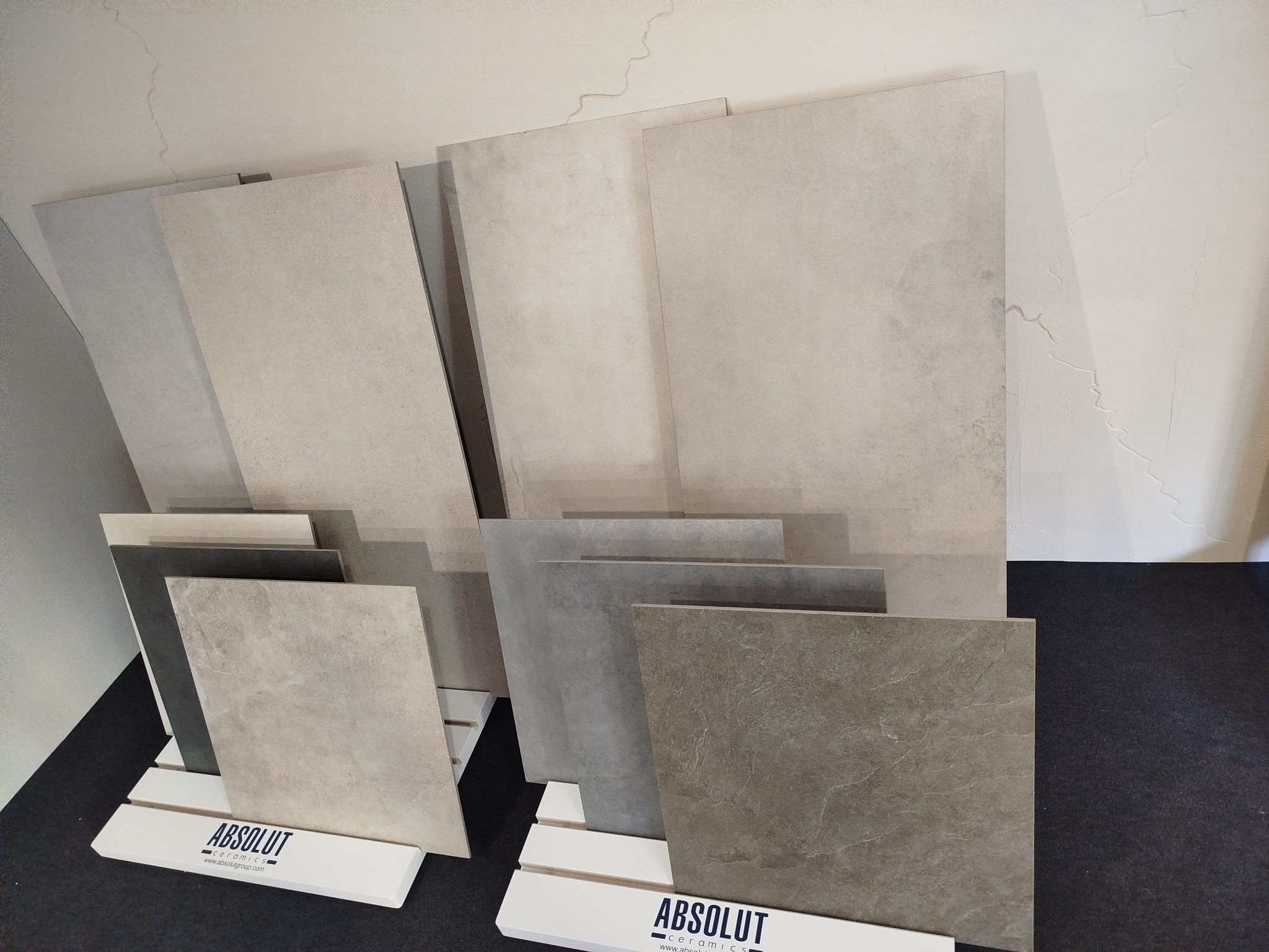 Ekspozycja płytek ceramicznych w różnych odcieniach szarości i beżu, prezentowanych na stojakach z logo producenta, na tle jasnej ściany z delikatnymi pęknięciami.