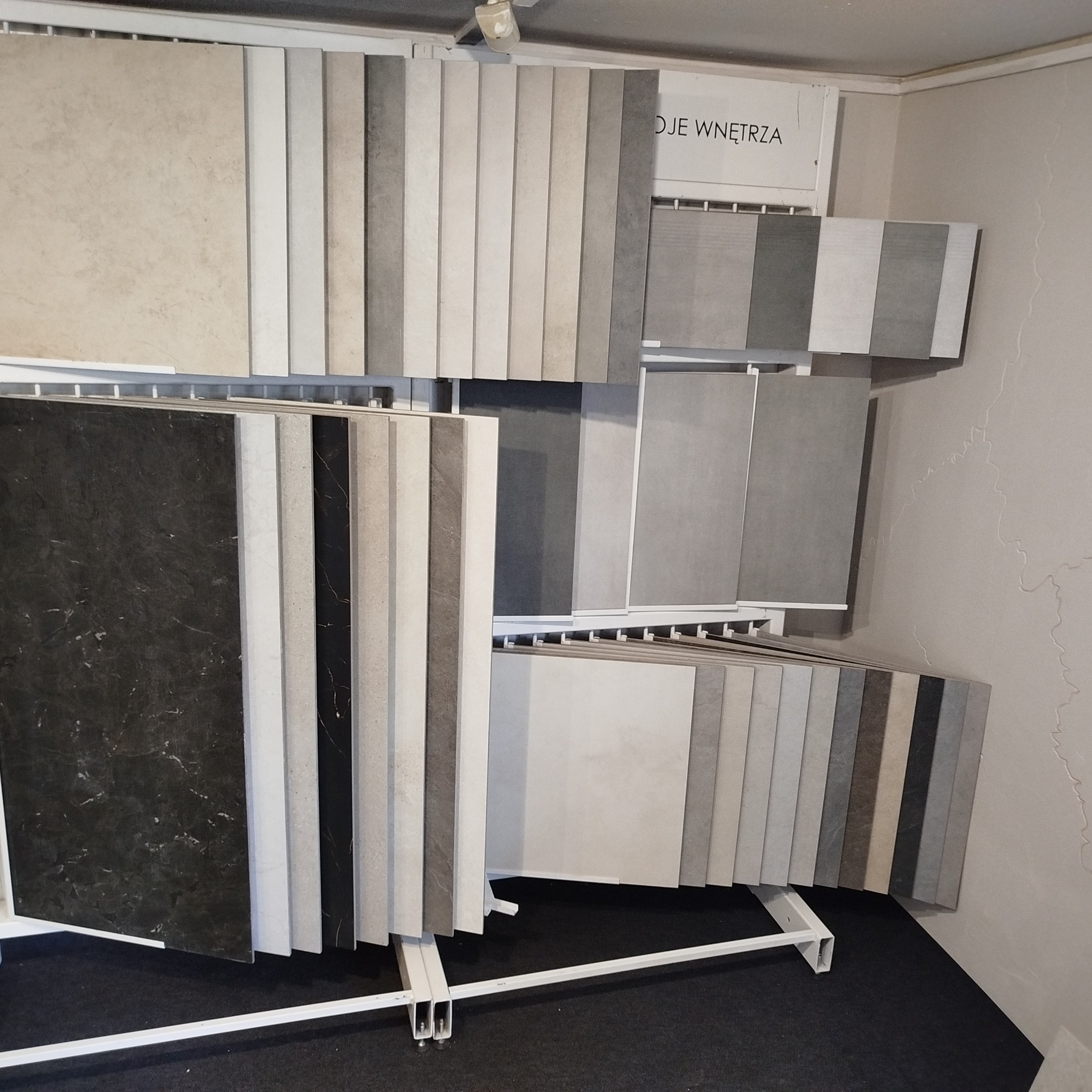 Ekspozycja różnorodnych płytek ceramicznych w odcieniach szarości, beżu i czerni, umieszczonych na metalowym stojaku w salonie z materiałami wykończeniowymi.