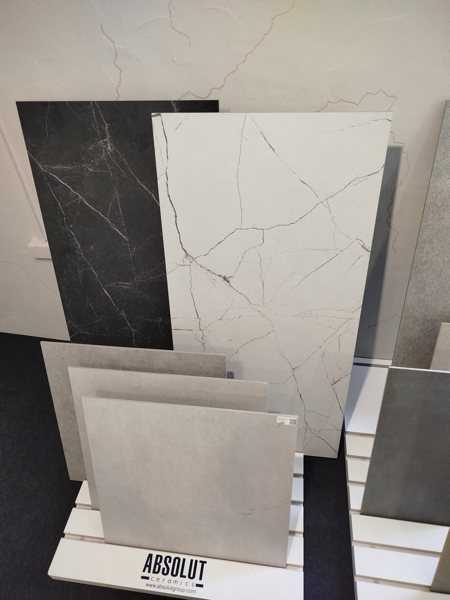 Ekspozycja płytek ceramicznych w różnych odcieniach szarości i bieli, z wzorem marmuru, ustawionych na białym stojaku z logo producenta.