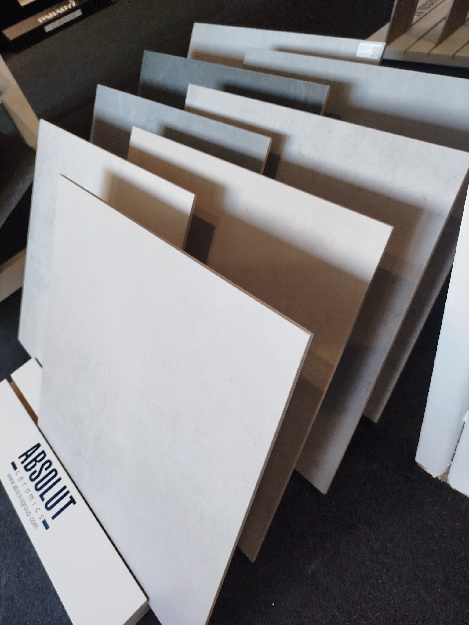 Ekspozycja płytek ceramicznych w odcieniach szarości i bieli, ułożonych warstwowo pod kątem, z widocznym logo producenta 'Absolut Ceramics'.