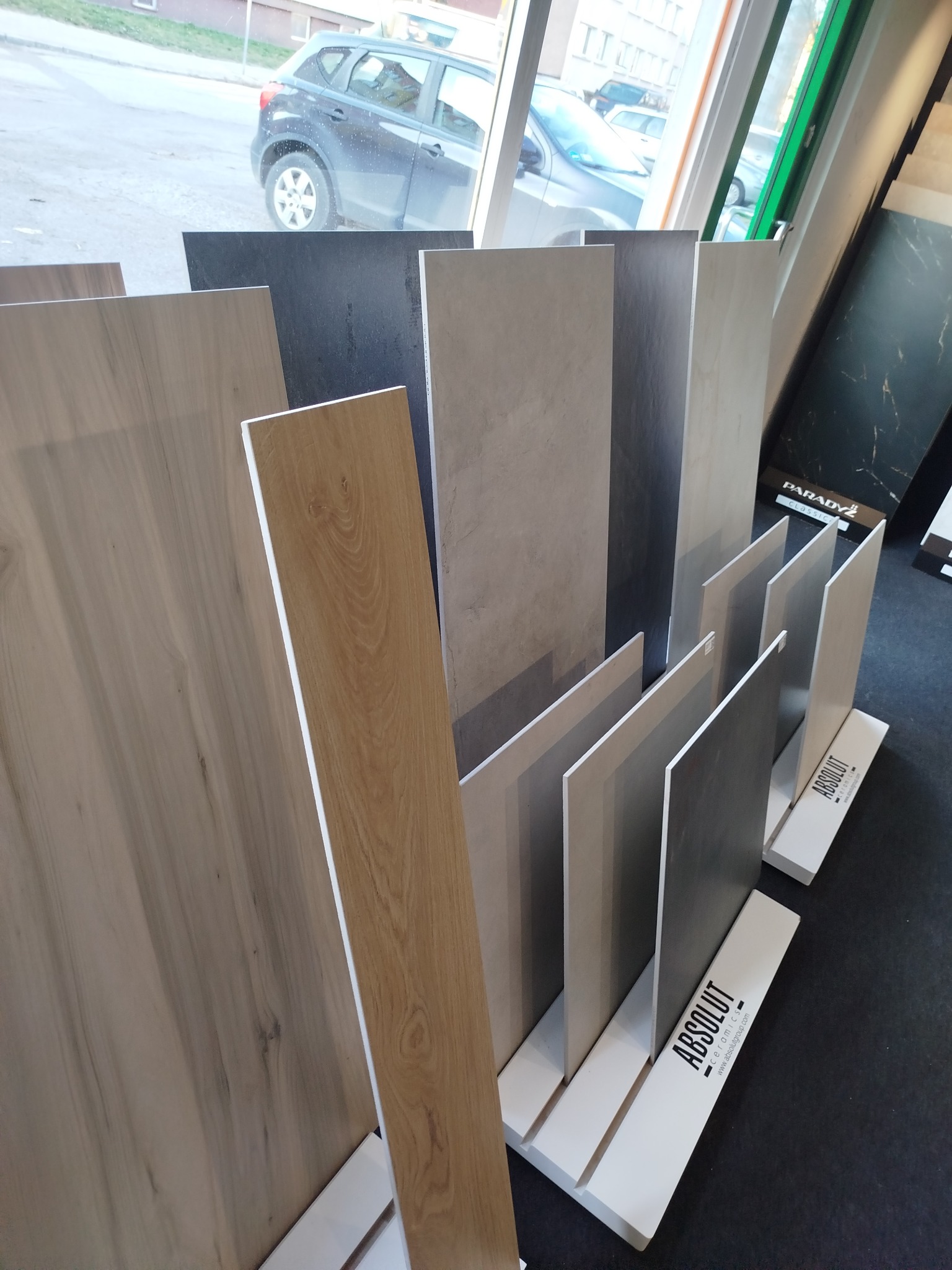 Ekspozycja różnorodnych płytek ceramicznych w salonie, prezentująca wzory imitujące drewno i kamień, ustawione na stojakach z logo producenta.