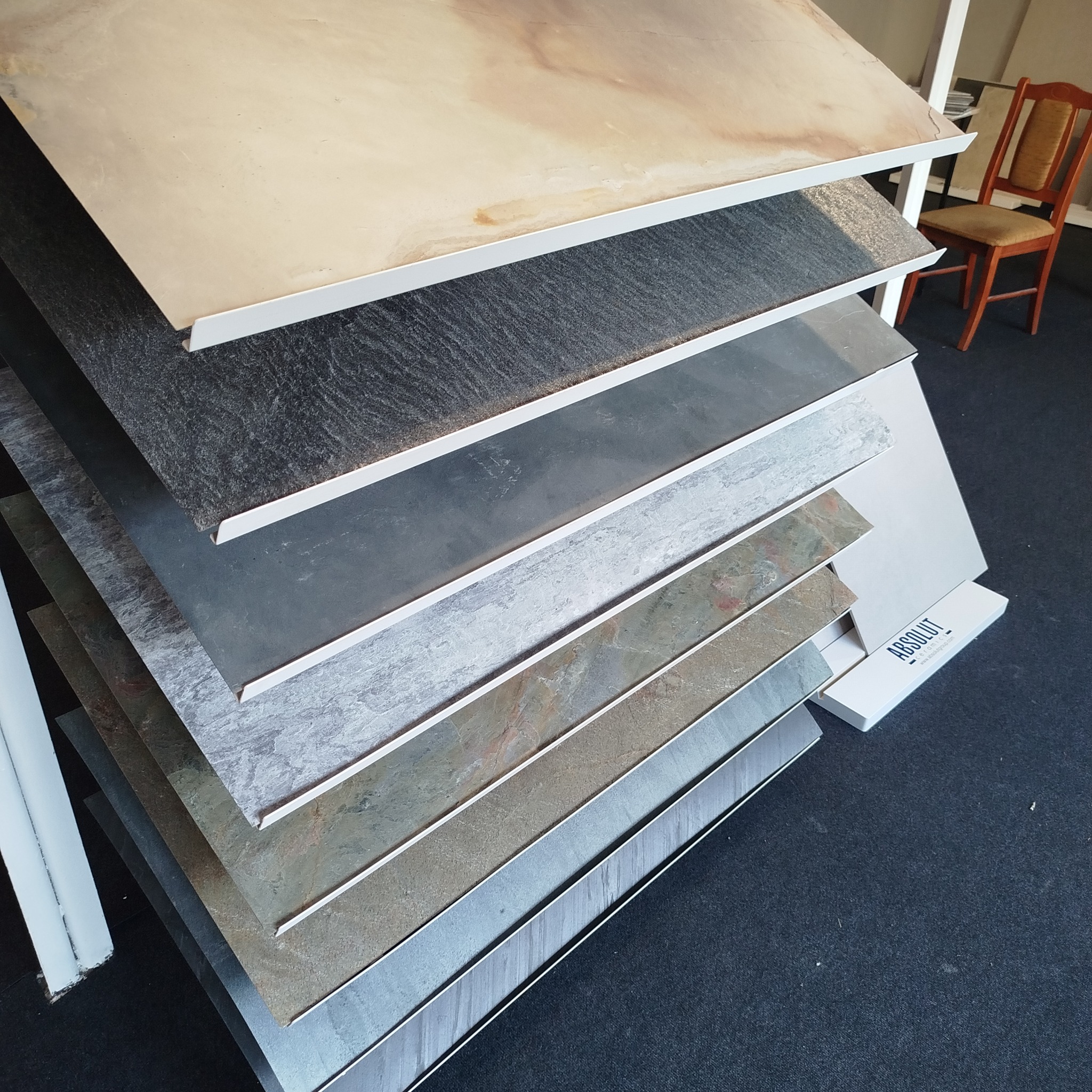 Ekspozycja próbek płytek ceramicznych o różnych wzorach i kolorach, ułożonych warstwowo na stojaku. W tle widoczne krzesło.