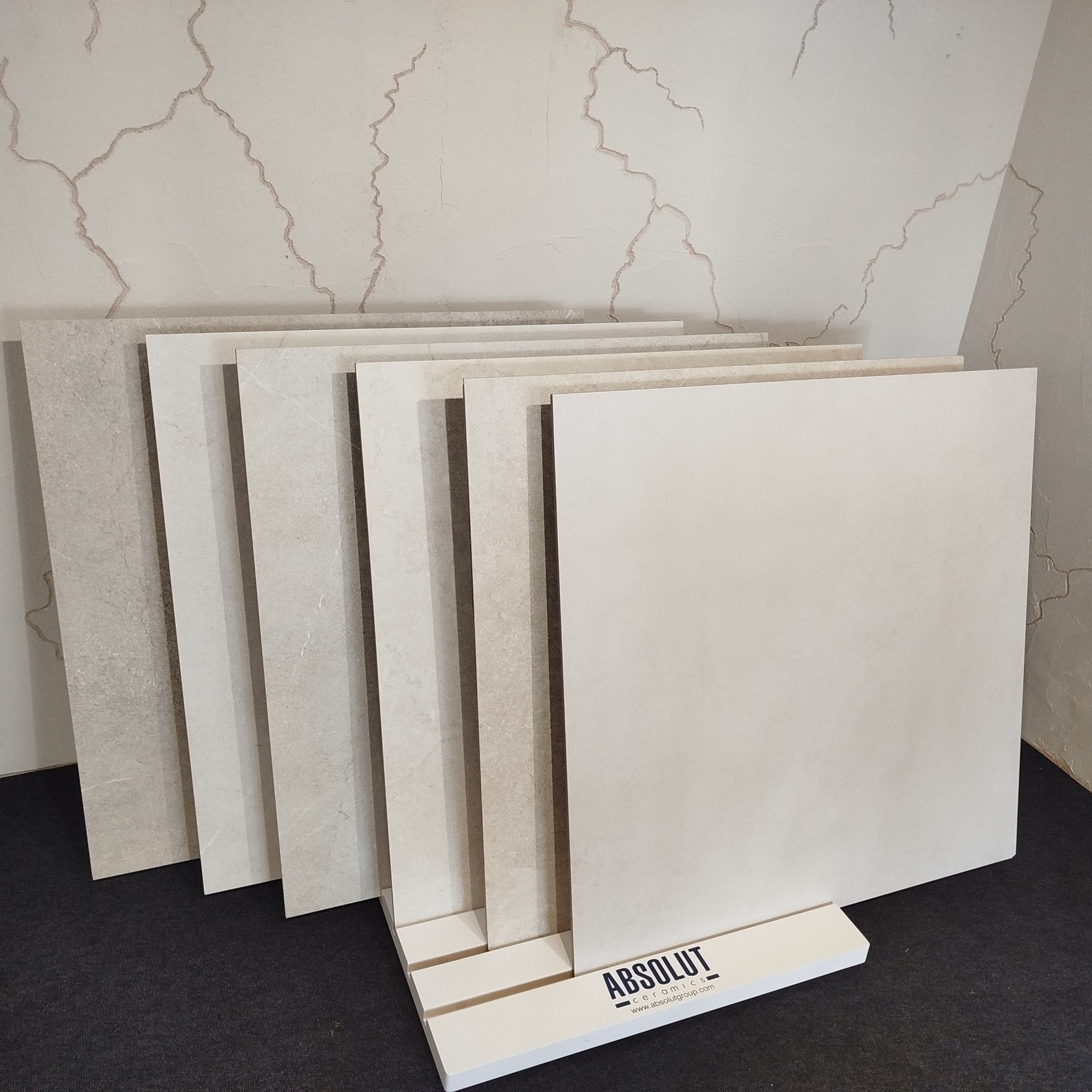 Ekspozycja płytek ceramicznych w odcieniach beżu, ustawionych na stojaku z logo producenta, na tle ściany z delikatnym wzorem imitującym spękania.