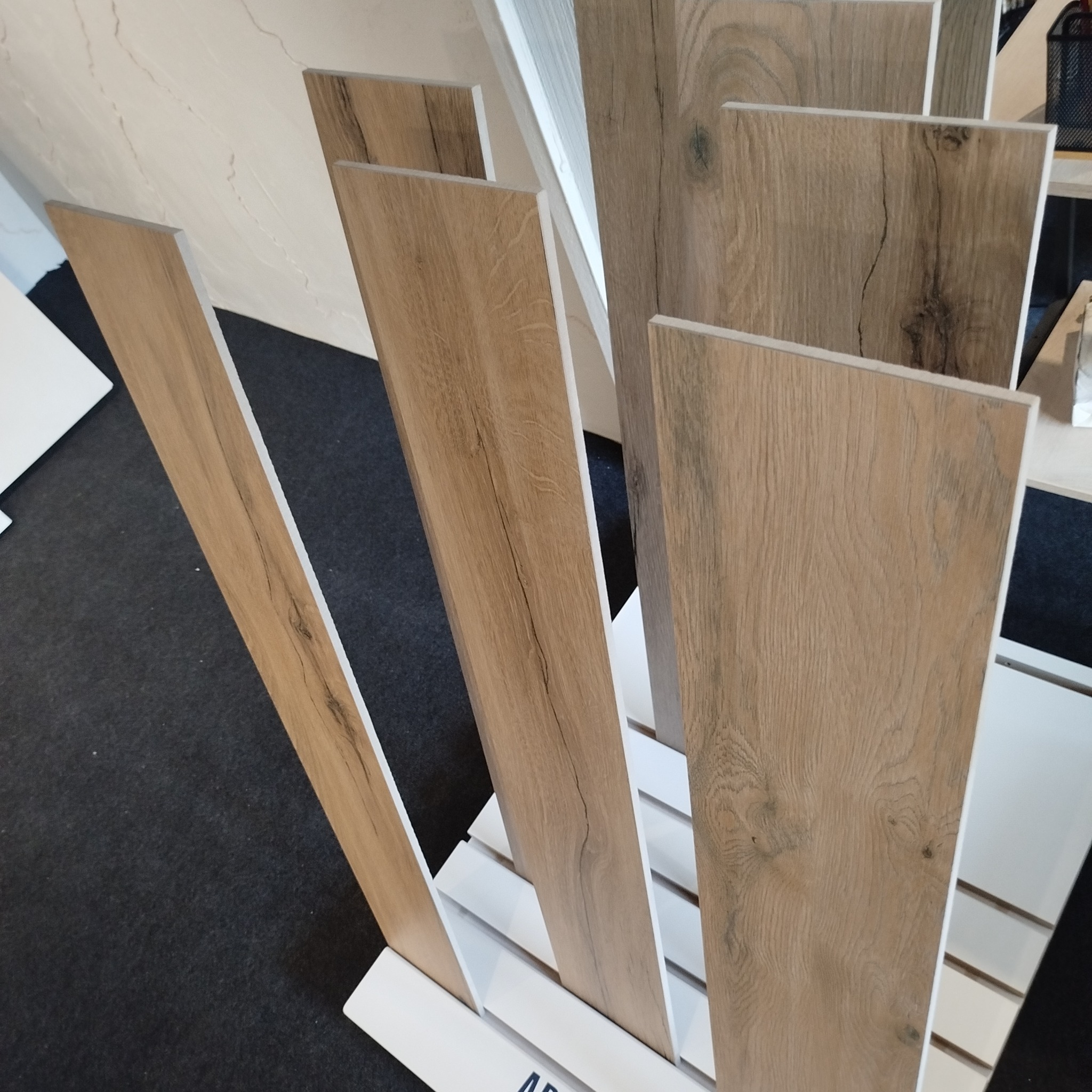 Ekspozycja płytek podłogowych imitujących drewno, ustawionych pionowo na białym stojaku. Widoczne różne odcienie i wzory drewna, z bliska ukazujące strukturę.