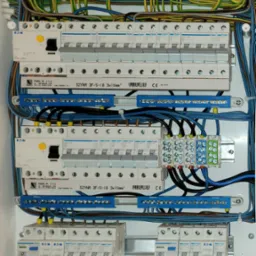 Rozdzielnica elektryczna z zabezpieczeniami różnicowoprądowymi i nadprądowymi firm Hager i Eaton, okablowana przewodami w kolorach żółto-zielonym, niebieskim i czarnym, z szynami zbiorczymi...