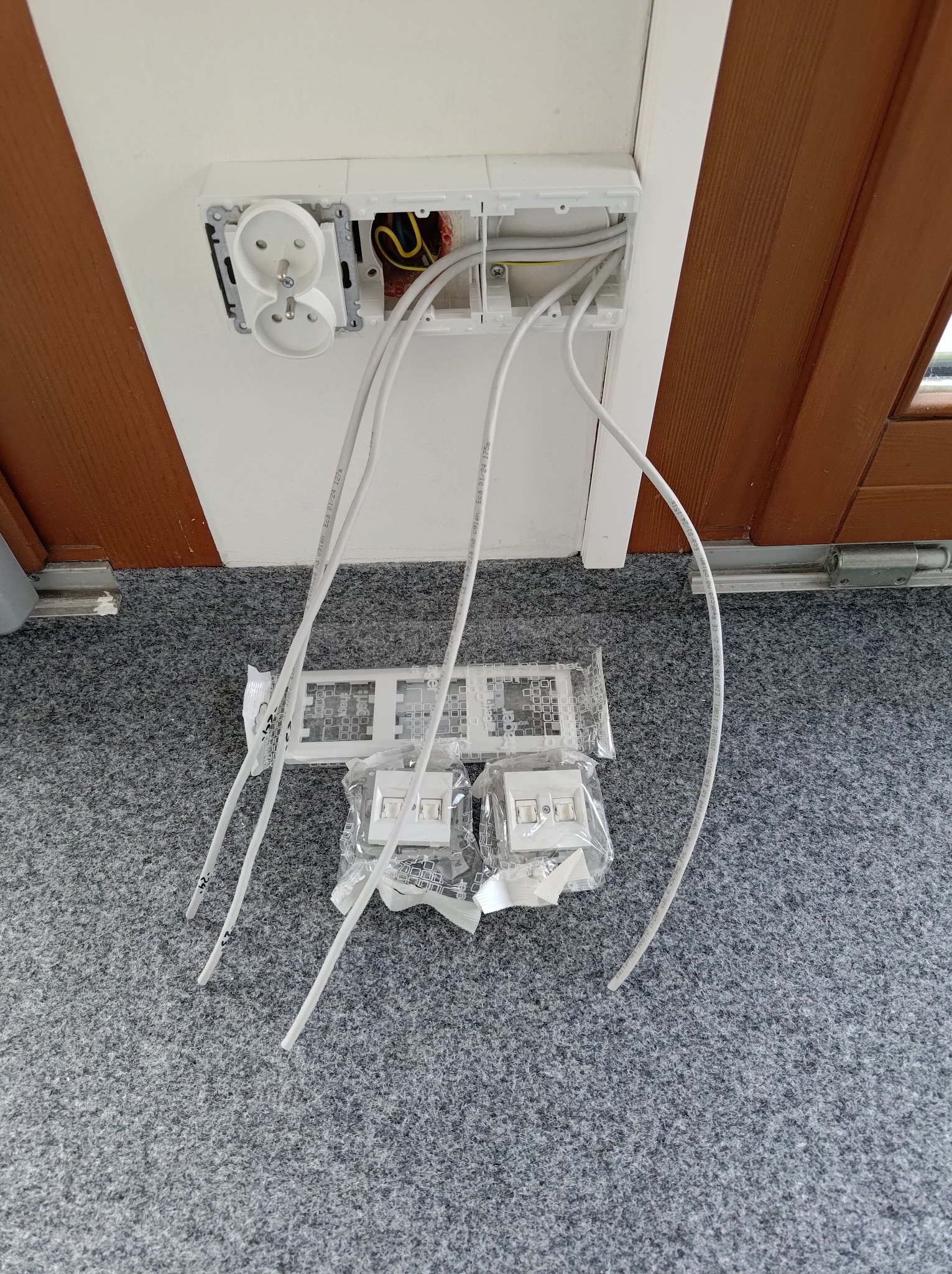 Podwójne gniazdko elektryczne z odsłoniętymi przewodami w puszce, obok leżą nowe, zapakowane elementy instalacji, na szarym dywanie.