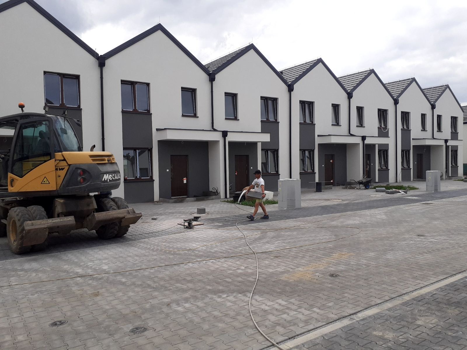 Nowe, szeregowe domy z szaro-białą elewacją, widoczny pracownik z wężem, maszyna budowlana i kostka brukowa na podjeździe, prawdopodobnie w trakcie prac wykończeniowych.
