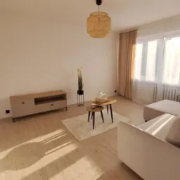 Jasne panele podłogowe w salonie po remoncie, z widoczną szafką RTV, stolikami kawowymi i beżową sofą, oświetlone popołudniowym światłem.