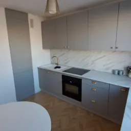 Nowoczesna kuchnia w odcieniach szarości z marmurowym blatem i złotymi akcentami, widok na okno z owocami i parkiet w jodełkę.