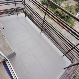 VITALII DERDIIENKO VIDE-REM - Wykończony balkon z szarymi płytkami, widok z góry przez metalową balustradę. W tle budynki i zieleń. Widoczny fragment buta w lewym dolnym rogu.