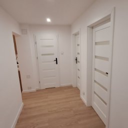VITALII DERDIIENKO VIDE-REM - Jasny korytarz po remoncie: panele podłogowe imitujące drewno, białe ściany i trzy białe drzwi z czarnymi klamkami. Minimalistyczny design, oświetlenie punktowe.