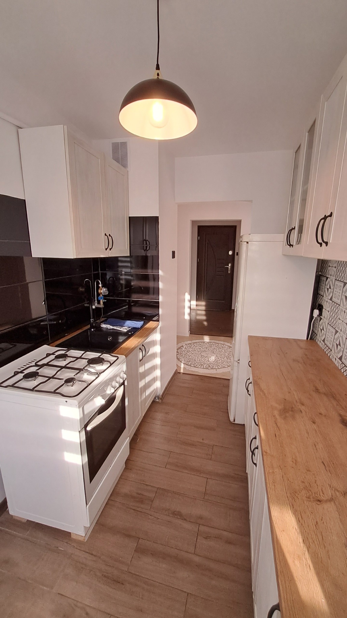 Wąska kuchnia z białymi szafkami, czarnym blatem i płytkami, drewnianą podłogą i wiszącą lampą w stylu retro, widok na przedpokój z okrągłym dywanem.