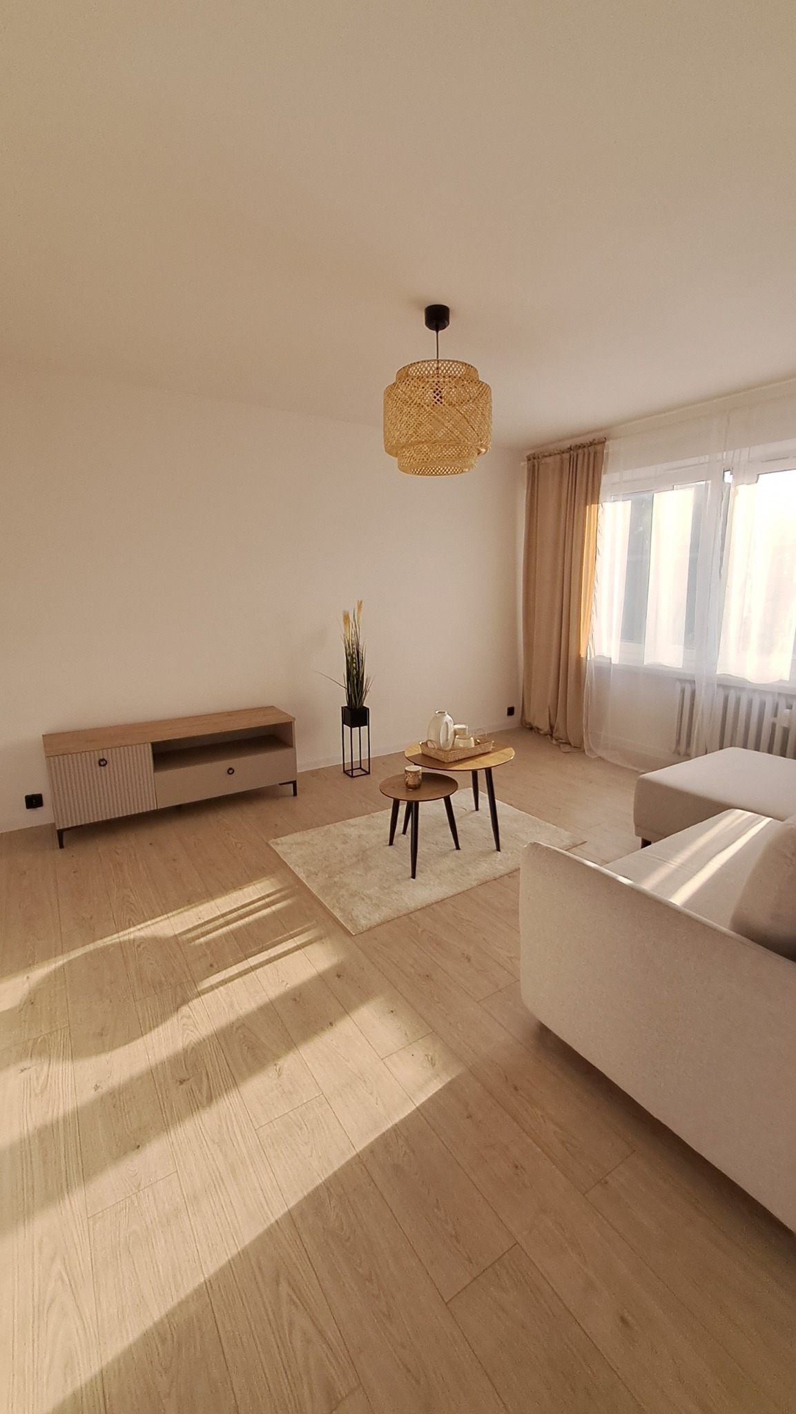 Jasne panele podłogowe w salonie po remoncie, z widoczną szafką RTV, stolikami kawowymi i beżową sofą, oświetlone popołudniowym światłem.