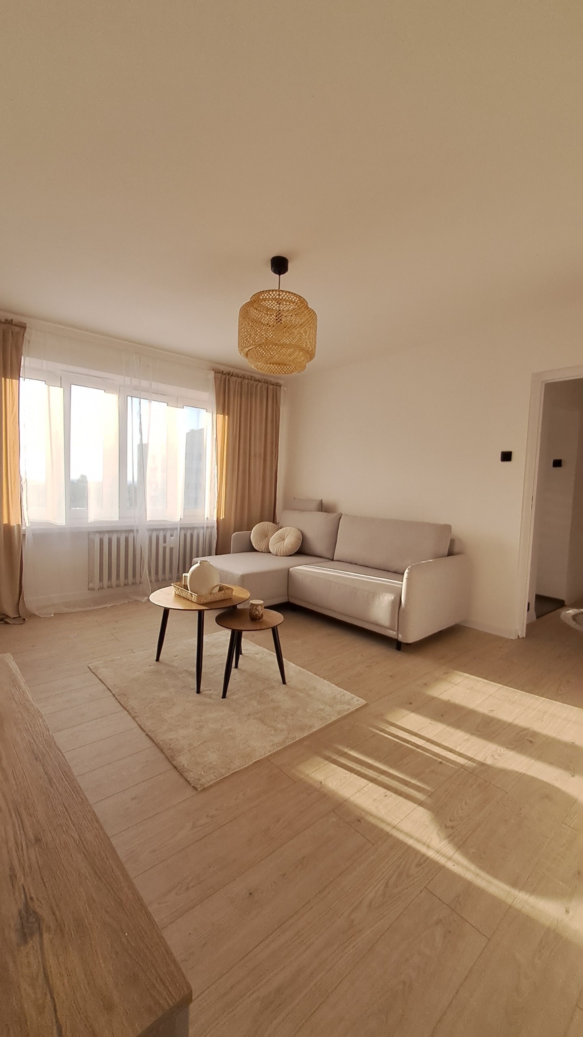 Jasny salon z beżową sofą narożną, okrągłymi stolikami kawowymi na dywanie i rattanową lampą wiszącą, parkiet w odcieniach drewna, promienie słoneczne wpadające przez okno.