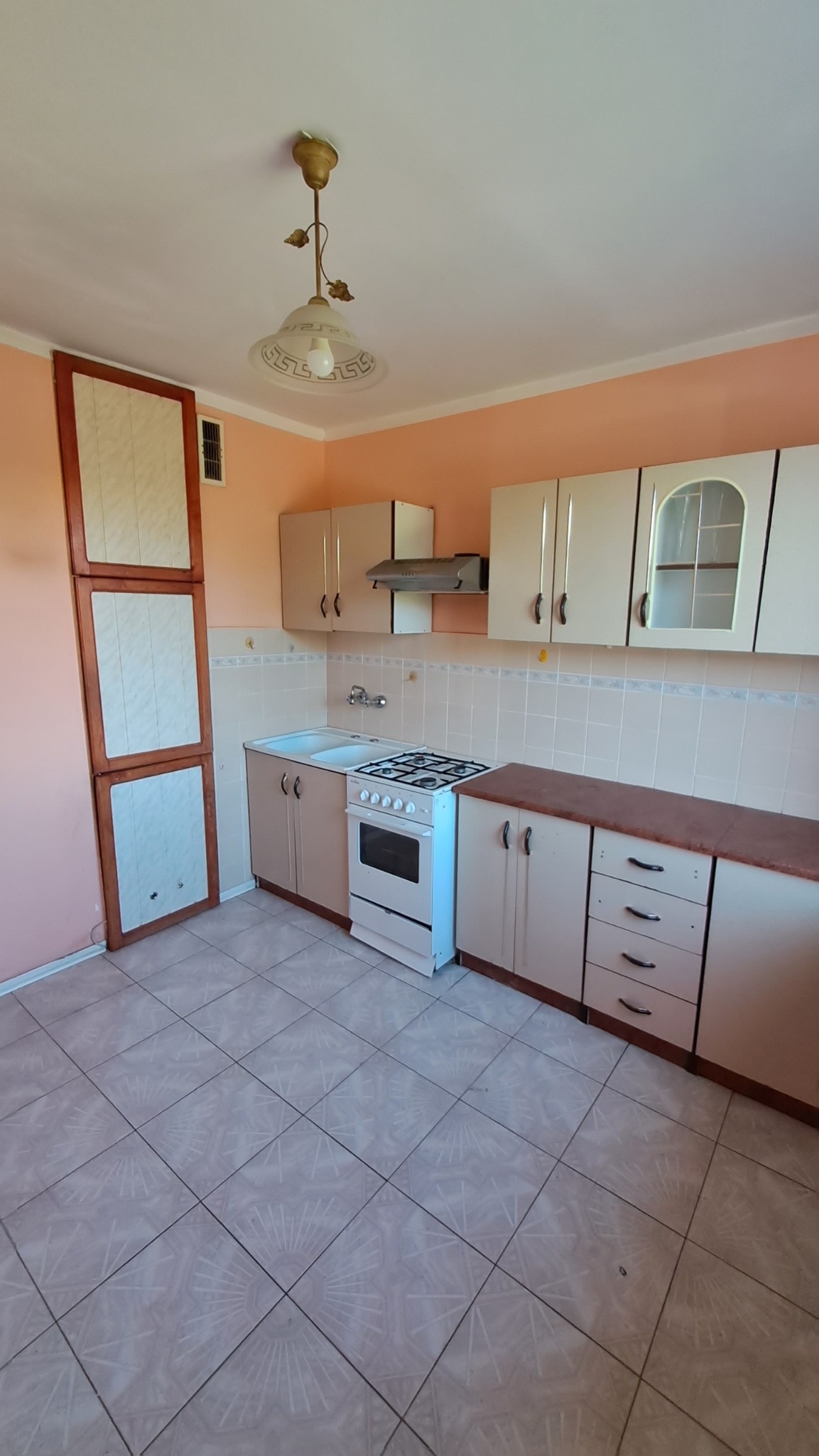 Jasna kuchnia z beżowymi szafkami, białym piecem i wzorzystą podłogą z płytek, sfotografowana z szerokiego kąta ukazującego całe pomieszczenie, w tym lampę sufitową z motywem roślinnym.