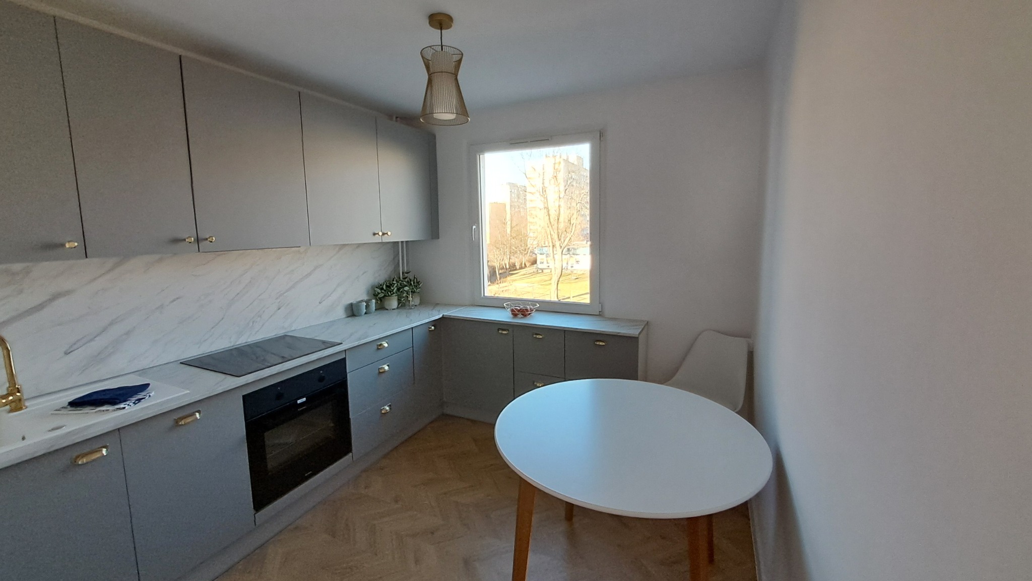 Jasna kuchnia po odnowieniu: szare szafki z marmurowym blatem i złotymi uchwytami, okrągły stół na drewnianych nogach, designerska lampa wisząca.