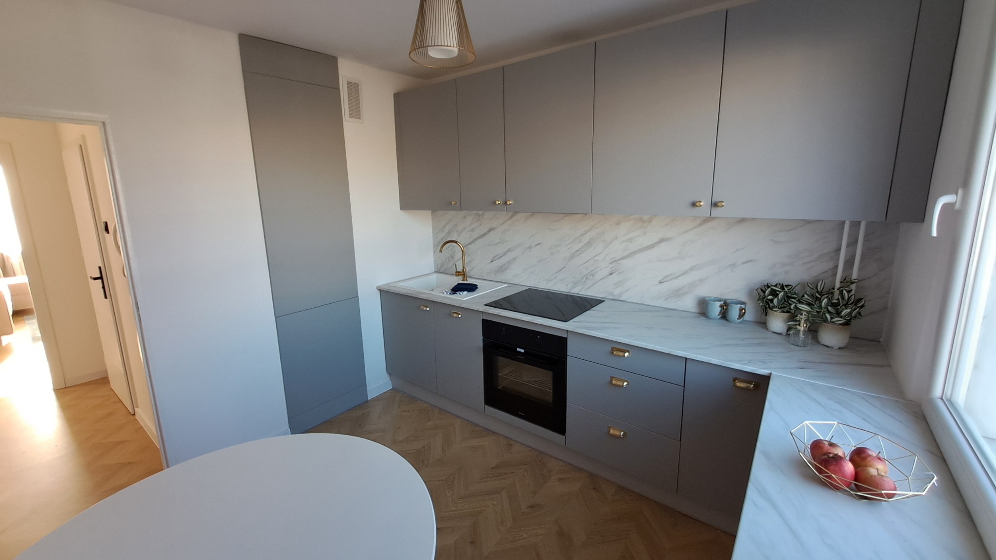 Nowoczesna kuchnia w odcieniach szarości z marmurowym blatem i złotymi akcentami, widok na okno z owocami i parkiet w jodełkę.