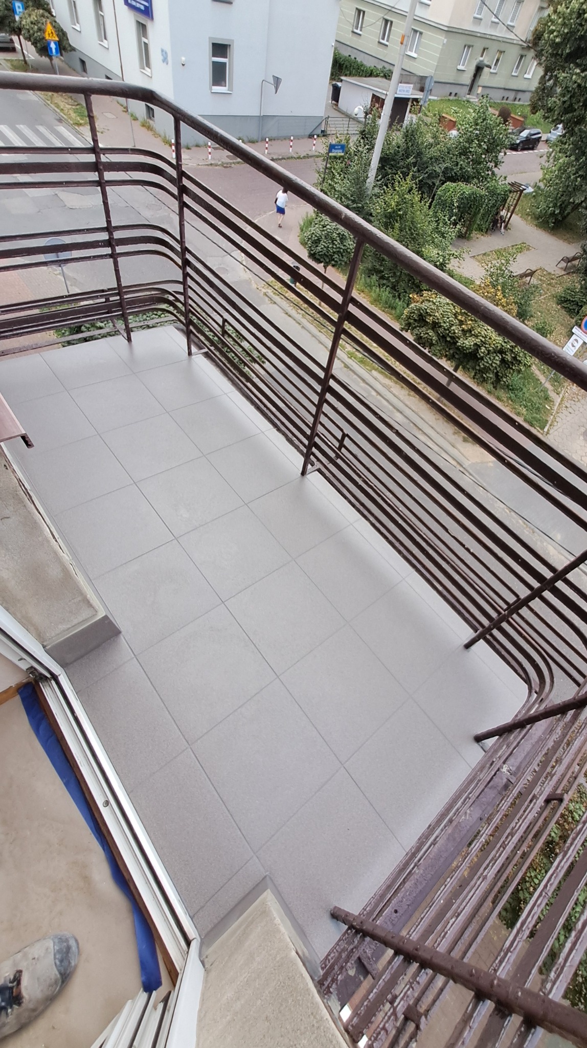 Wykończony balkon z szarymi płytkami, widok z góry przez metalową balustradę. W tle budynki i zieleń. Widoczny fragment buta w lewym dolnym rogu.