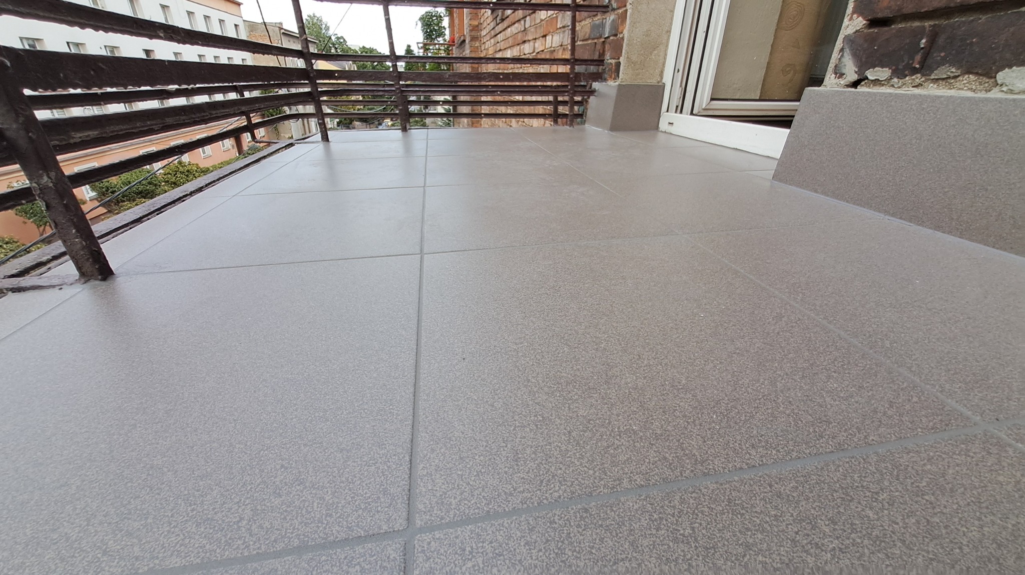 Wykończony balkon z szarymi, kwadratowymi płytkami. Widoczne barierki balkonu i fragment budynku w tle. Ujęcie z niskiej perspektywy podkreśla wzór płytek.