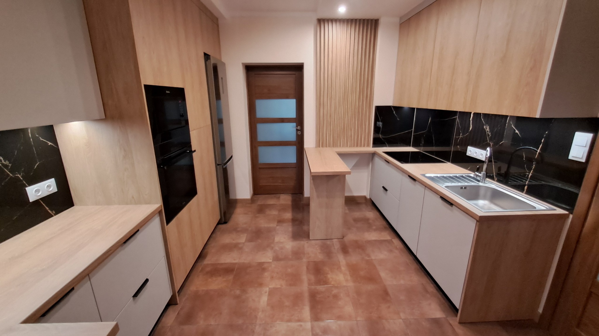 Nowoczesna kuchnia po remoncie: jasne szafki, blat imitujący drewno, czarny panel z marmurowym wzorem. Widoczna zabudowa i brązowe płytki podłogowe.