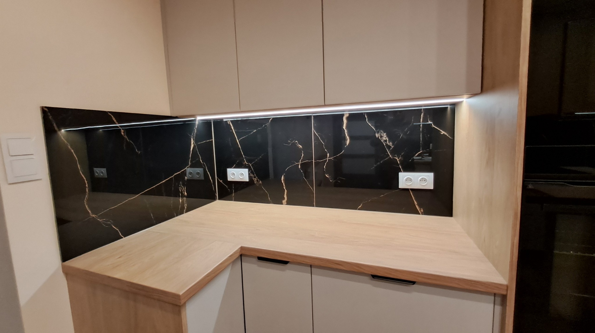 Nowoczesna kuchnia z czarnym panelem ściennym imitującym marmur ze złotymi żyłami, oświetleniem LED podszafkowym i drewnianym blatem.