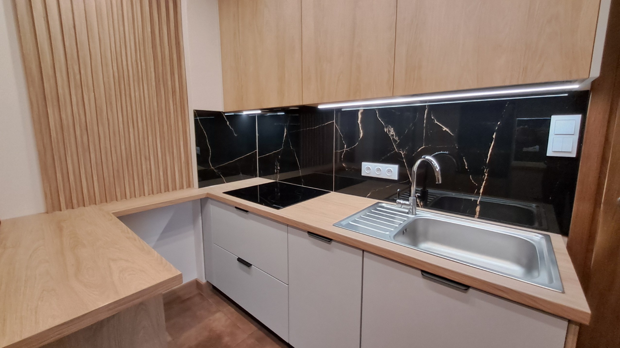 Nowoczesna kuchnia z drewnianymi szafkami, czarnym blatem z żyłami i panelem dekoracyjnym. Ujęcie z bliska na zlew i płytę indukcyjną.