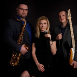 Trzyosobowy zespół muzyczny w studiu: saksofonista w okularach, wokalistka z mikrofonem i gitarzysta w szarej marynarce, na czarnym tle.