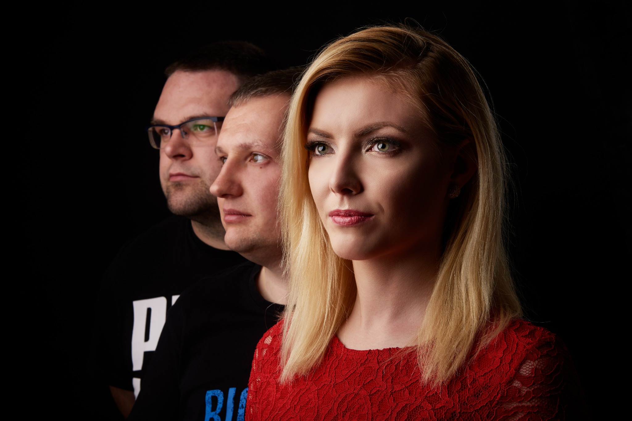 Portret studyjny: troje muzyków ustawionych w rzędzie, z przodu blondwłosa wokalistka w czerwonej koronkowej sukience, za nią dwaj mężczyźni w czarnych koszulkach z białymi i niebieskimi napisami...