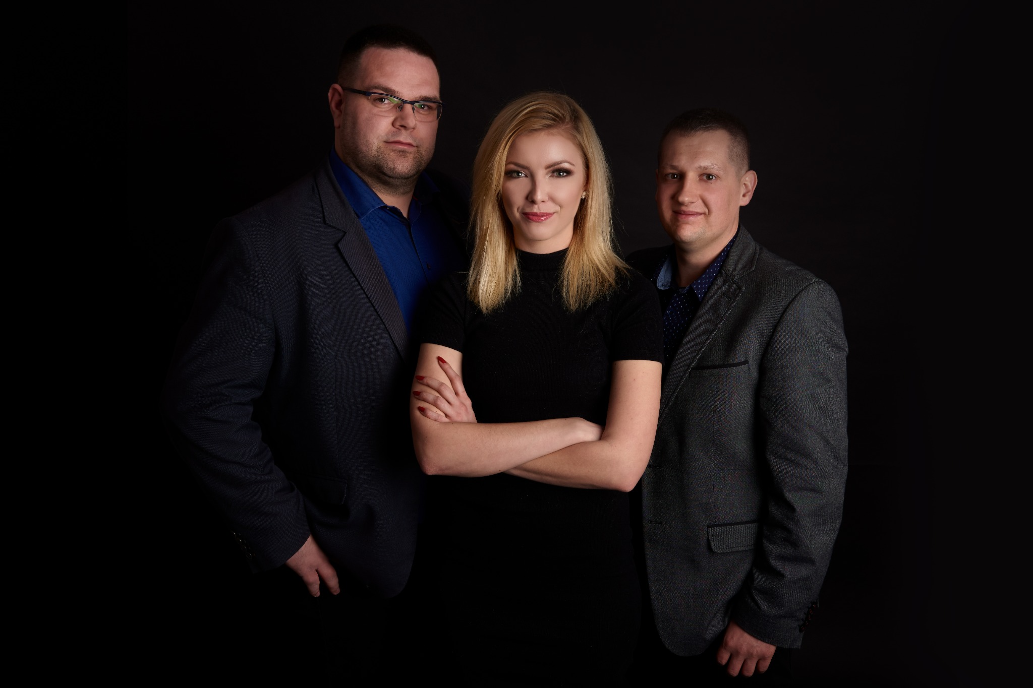 Profesjonalny portret studyjny: kobieta z blond włosami, skrzyżowanymi ramionami, w czarnej sukience, otoczona dwoma mężczyznami w marynarkach, na czarnym tle.