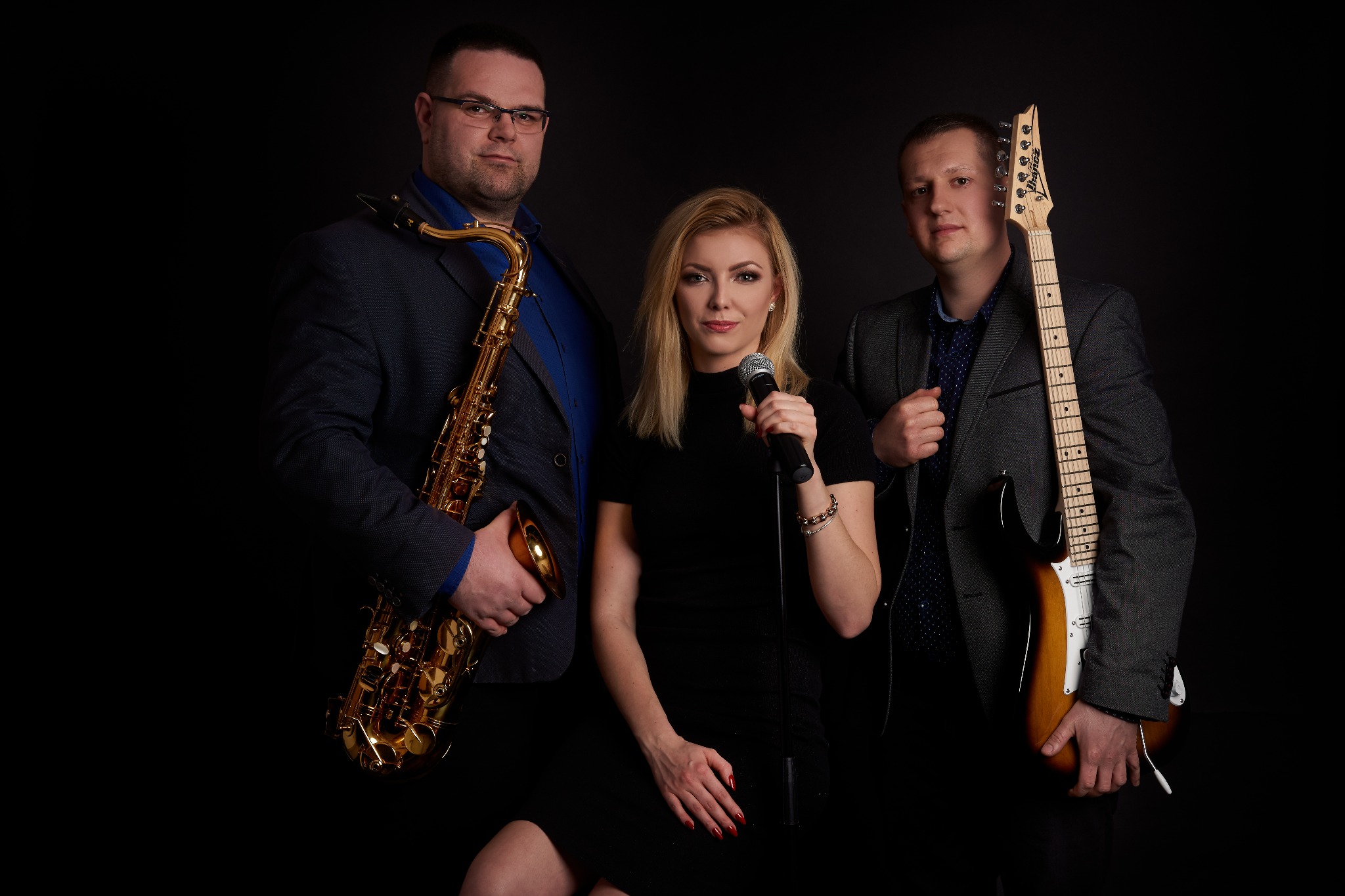 Trzyosobowy zespół muzyczny w studiu: saksofonista w okularach, wokalistka z mikrofonem i gitarzysta w szarej marynarce, na czarnym tle.