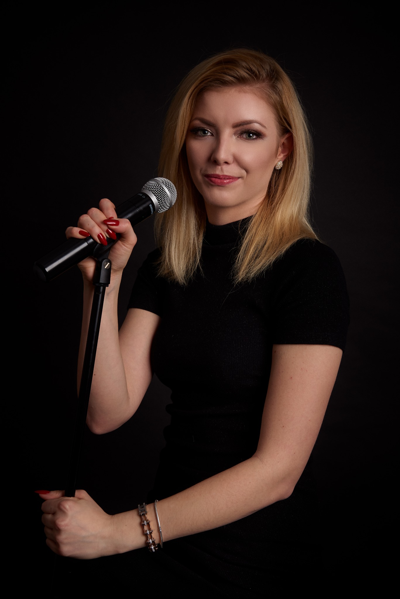 Portret uśmiechniętej blondynki z mikrofonem Shure na czarnym tle, z widocznymi czerwonymi paznokciami i srebrną bransoletką.