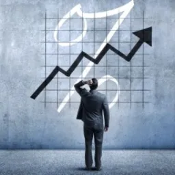 Mężczyzna w garniturze stoi przed ścianą z namalowanym symbolem procentu i strzałką skierowaną w górę, zamyślony.