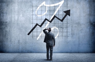 Mężczyzna w garniturze stoi przed ścianą z namalowanym symbolem procentu i strzałką skierowaną w górę, zamyślony.