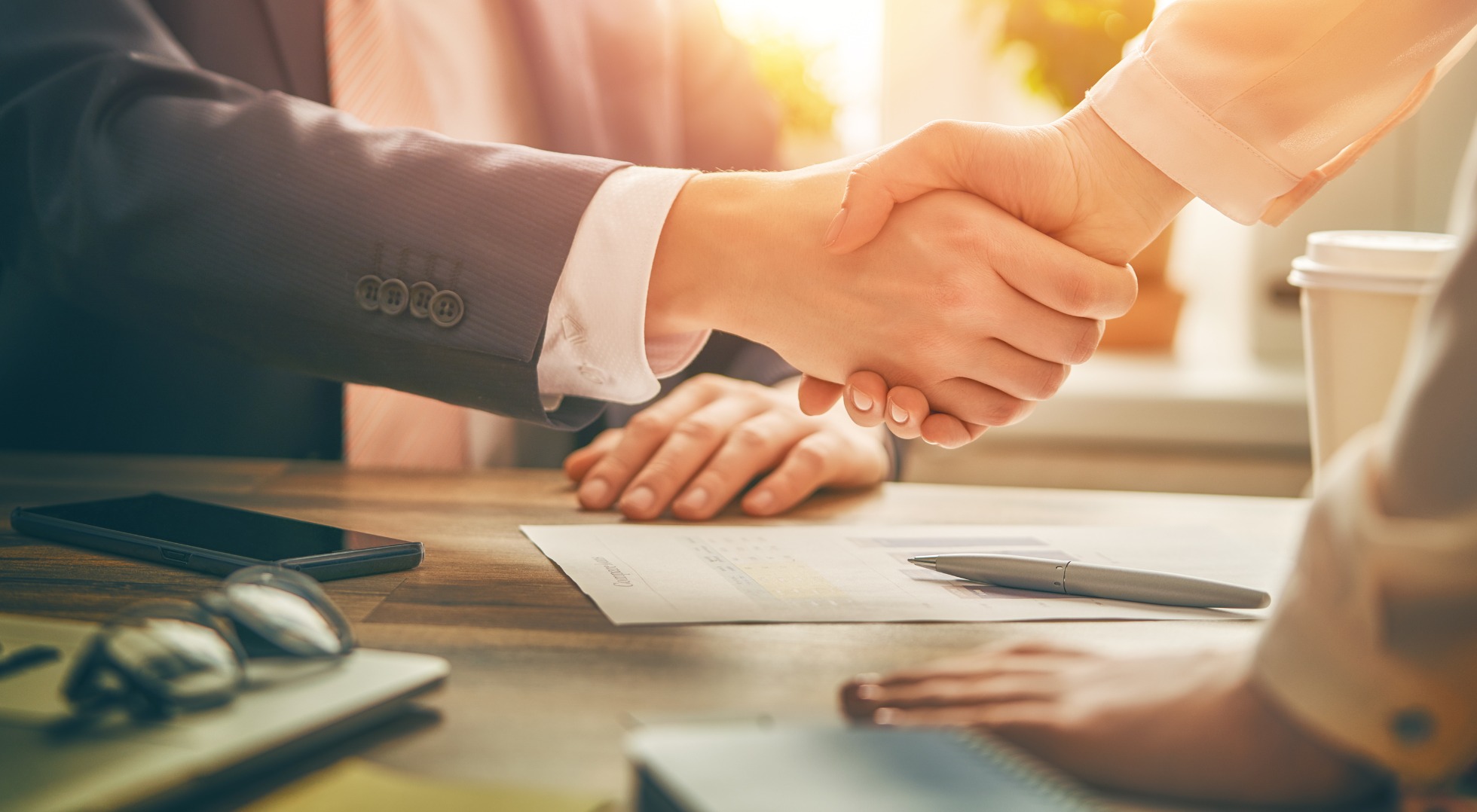 Uścisk dłoni nad dokumentem z tabelami na drewnianym biurku, obok leżą okulary, smartfon i notatnik, w tle kubek z kawą i blask słońca.