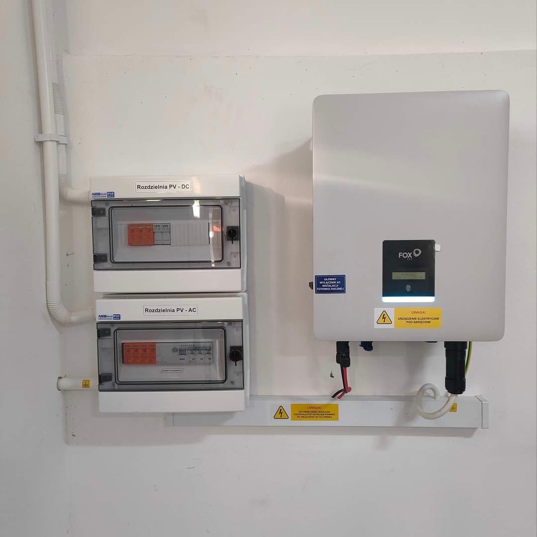 Biała skrzynka inwertera fotowoltaicznego FoxEss zamontowana na ścianie obok dwóch rozdzielnic PV-DC i PV-AC z widocznymi bezpiecznikami i oznaczeniami, z naklejkami ostrzegawczymi o napięciu...