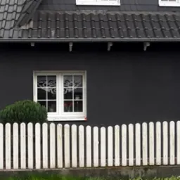 Dom z ciemną elewacją, białymi oknami z dekoracyjnymi naklejkami i białą, drewnianą sztachetową barierką. W tle widoczne rusztowanie i fragment boiska sportowego.