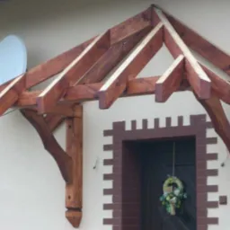 Drewniana wiata nad drzwiami wejściowymi z dekoracyjnym wieńcem, wsparta na rzeźbionych słupach, na tle jasnej elewacji z anteną satelitarną.