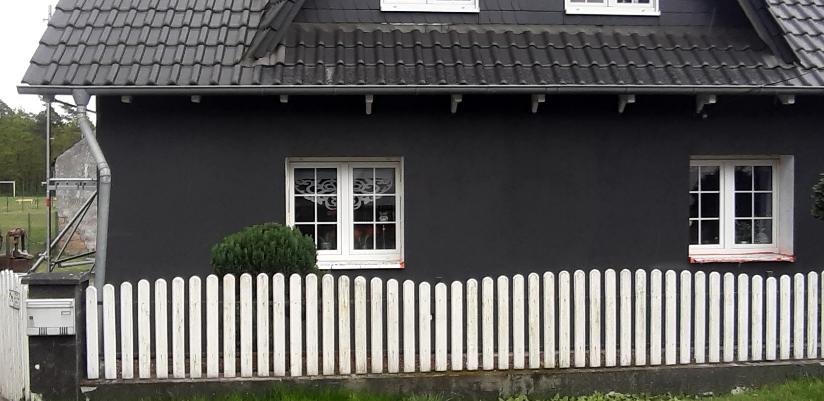 Dom z ciemną elewacją, białymi oknami z dekoracyjnymi naklejkami i białą, drewnianą sztachetową barierką. W tle widoczne rusztowanie i fragment boiska sportowego.