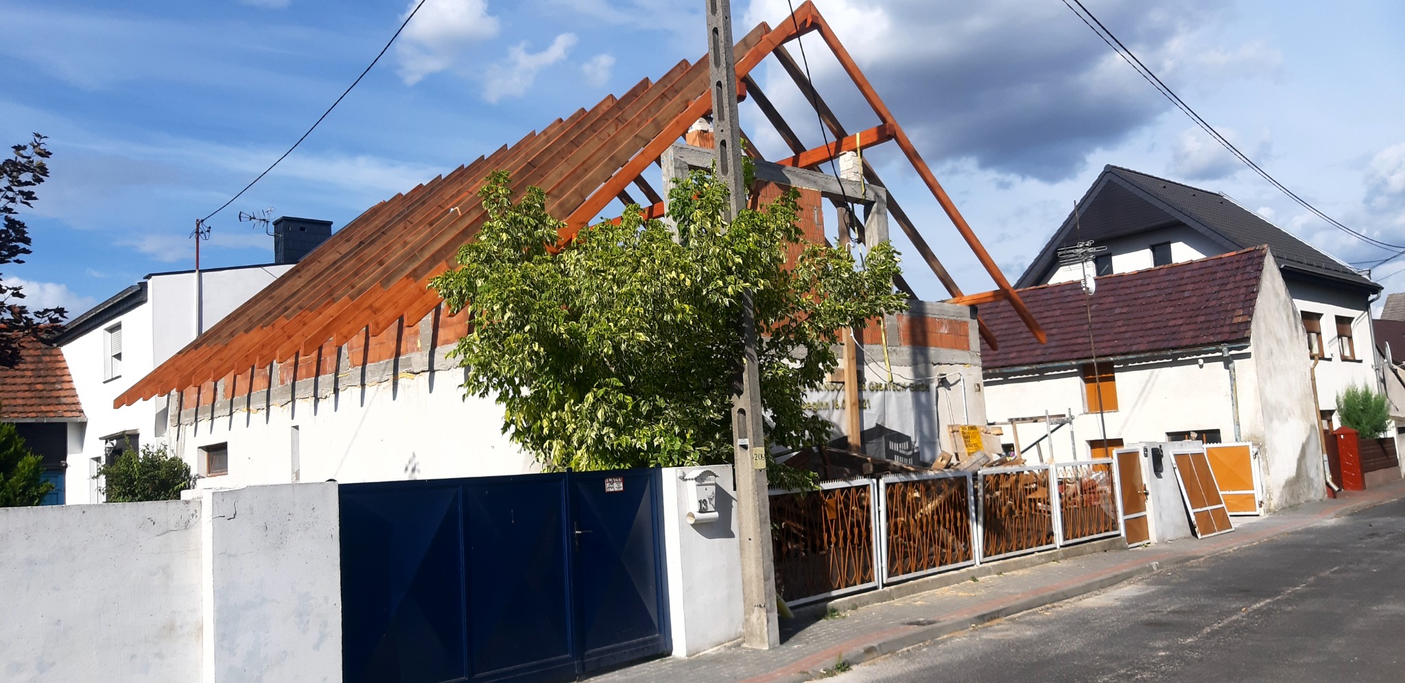 Konstrukcja drewniana dachu w trakcie budowy, widoczne krokwie i murłaty, z boku drzewo i słup energetyczny, w tle domy sąsiadów.