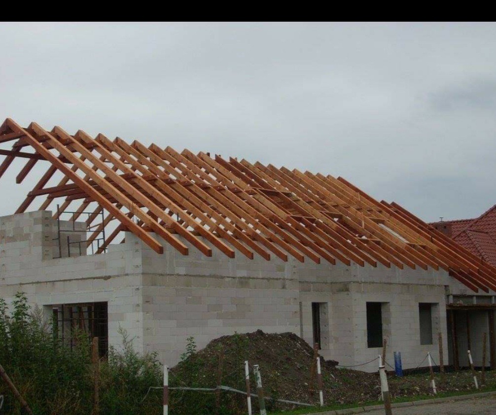 Konstrukcja drewniana dachu w trakcie budowy domu z białych bloczków, widoczne krokwie pod kątem na tle pochmurnego nieba.