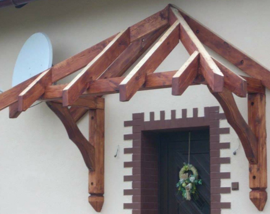 Drewniana wiata nad drzwiami wejściowymi z dekoracyjnym wieńcem, wsparta na rzeźbionych słupach, na tle jasnej elewacji z anteną satelitarną.