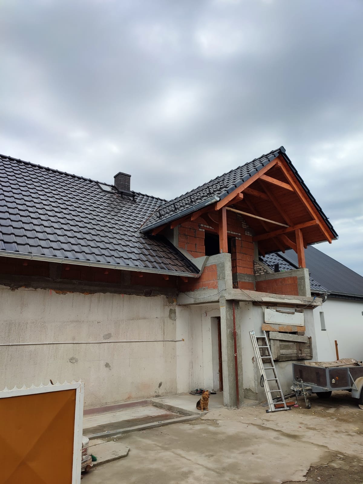 Dom w trakcie budowy z nowym, ciemnym dachem z dachówki ceramicznej, drewnianą konstrukcją werandy i niezakończonymi ścianami zewnętrznymi; pies siedzi na podwórku.